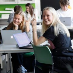 To elever sidder ved computer. Den ene rækker hånden i vejret.